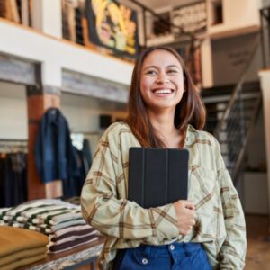 a smiling young woman holding a digital tablet stands in front of a clothing display in a retail store