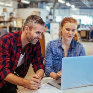 a smiling middle-aged man and a young woman in a commercial workshop look at a laptop computer